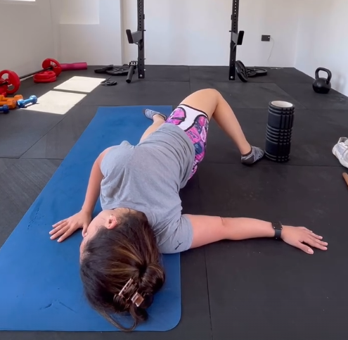 Jalu haciendo un estiramiento de espalda y piernas sobre un mat, con elementos de entrenamiento funcional de fondo